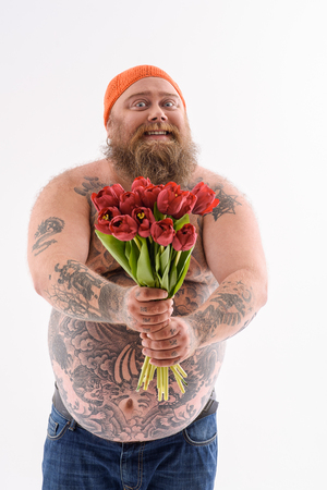 These flowers are for you. Joyful bearded fatty is holding tulips and stretching it to camera. Man smiling and looking forward with aspiration. Isolatesの写真素材