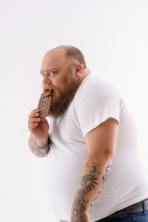 Hungry fat man is eating chocolate with desire. He is standing and looking forward with fear to be punished. Isolated on backgroundの写真素材