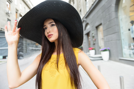 Stylish young woman is having walk in city. She is looking forward with interest. Lady is standing and touching hat elegantly Stylish young woman is having walk in city. She is looking forward with interest. Lady is standing and touching hat elegantly