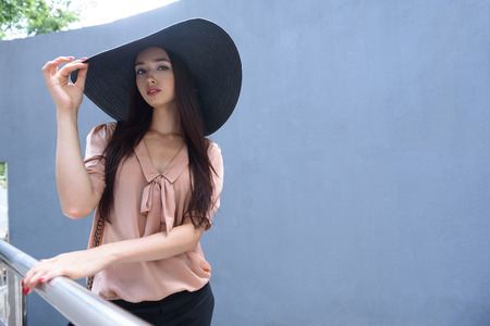 Elegant young woman is standing on balcony outdoors. She is touching her hat and looking at camera with confidenceの写真素材