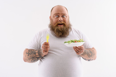 Joyful fat man is eating salad and smiling. He is standing and looking at camera with amazement....