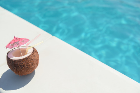 Close up of cocktail in coconut with straws and small umbrella near swimming poolの写真素材