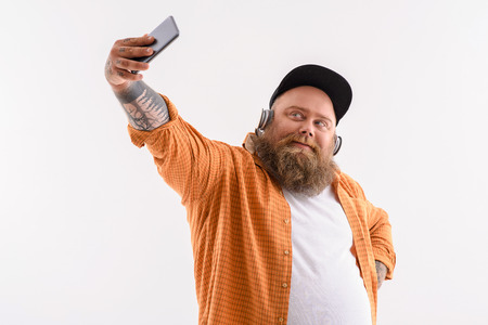 Joyful fat man is making selfie on smartphone. He is posing and smiling. Guy is wearing headphonesの写真素材