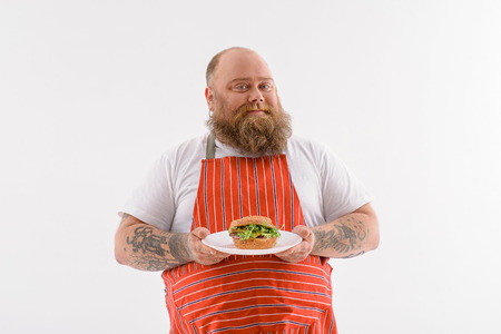 Happy fat man is holding plate with sandwich and showing it to camera. He is standing and smiling. Chef is wearing cooking apron. Isolatedの写真素材