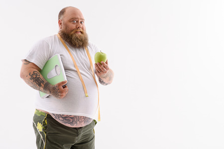 Fat man is dieting with joy. He is showing apple and smiling. Man is standing and holding scales with tape measure. Isolated and copy space in right sideの写真素材