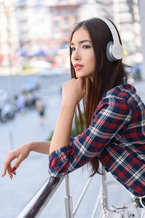 Beautiful young woman is enjoying music outdoors. She is looking at city on balcony and dreamingの写真素材