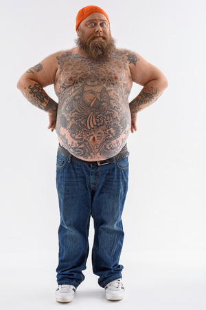 Fat man is standing with arms akimbo. He is looking at camera with surprise. Guy has big naked belly and beard. Isolatedの写真素材