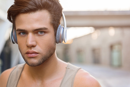 Attractive young man is listening to music from earphones. He is standing outdoors and looking...