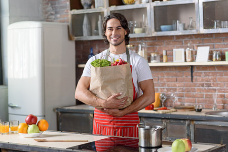 Happy young man is holding packet of healthy food. He is standing in kitchen and smilingの写真素材