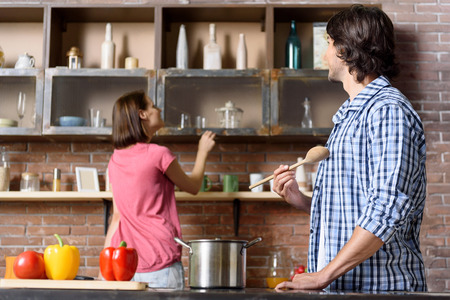 Joyful married couple is cooking together. Woman is looking for ingredient on shelf in kitchen. Man is standing near pot and holding wooden spoon. They are smilingの写真素材