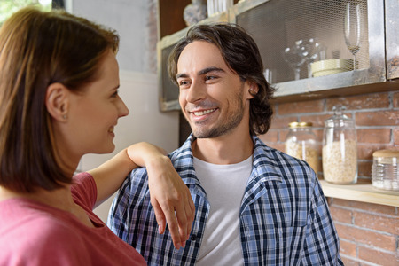 Happy young man and woman are looking at each other with love. They are talking and smiling. Lovers are standing in kitchenの写真素材