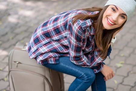 Carefree young girl is sitting on suitcase. She is looking at camera and smilingの写真素材