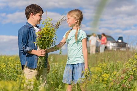 Very sweet moment. Little boy giving bouquet of wild flowers for beautiful girl, standing in field during travelの写真素材
