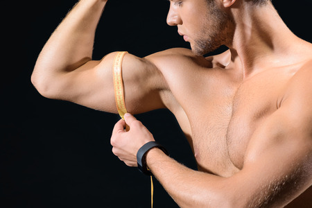 Professional young sportsman is measuring his bicep with tape. He is standing and looking at arm with concentration. Isolated on black backgroundの写真素材