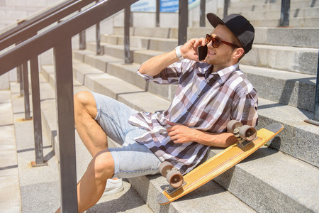 Cheerful male skater is talking on mobile phone and laughing. He is sitting on stairs outdoors and...