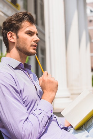 Talented young man is writing poem with inspiration. He is touching pencil to chin pensively. Guy is...