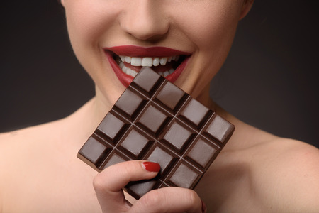 Close up of happy young woman eating chocolate with pleasure. She is standing and smiling