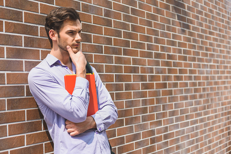 Thoughtful young man is standing near wall and holding folder. He is touching chin and looking forward pensivelyの写真素材