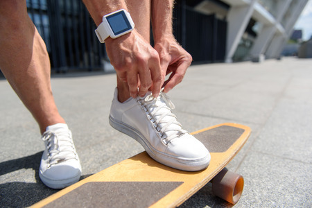Close up of male legs standing on skateboard. Man is tying laces on his sneakers. He has smart watch...