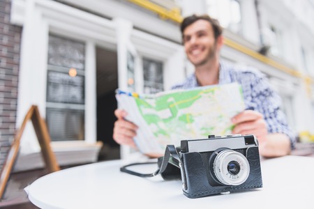 Inspired male tourist is planning route of his trip. He is sitting and holding map. Man is smiling. Focus on camera on tableの写真素材