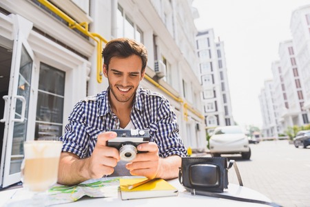 Happy young man is watching photos from trip. He is holding camera and smiling. Traveler is sitting at table in caf? outdoorsの写真素材