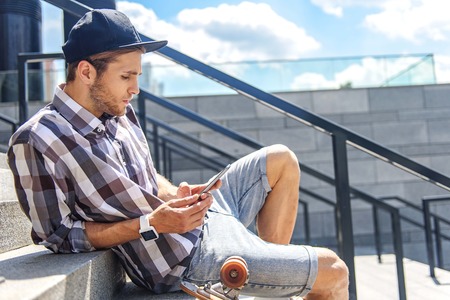 Cool young man is messaging on mobile phone with concentration. He is sitting on stairs near skate...