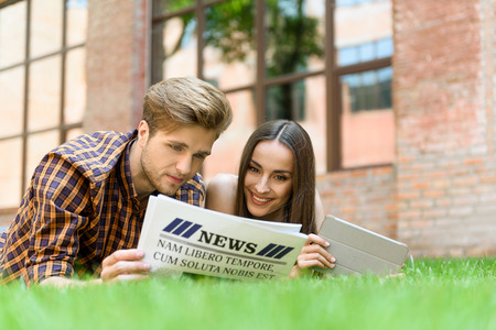 Happy friends are reading newspaper together. They are lying on lawn near building and laughing. Woman is holding tabletの写真素材