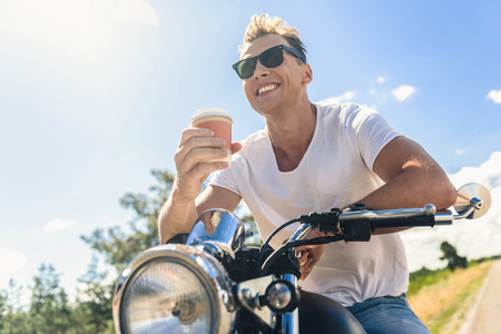 Time for coffee. Happy smiling guy sitting on his motorcycle and holding cup of coffee against blue skyの写真素材