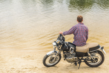 Thinking about something important. Rear view of young man sitting on his vintage motorcycle and looking away of lakeの写真素材