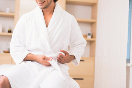 Joyful young man is having rest after bathing at spa. He is sitting and tying belt of bathrobe. Man is smiling happilyの写真素材