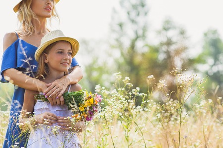 Carefree mother and daughter are standing and relaxing on meadow. They are embracing and smiling. Woman is holding flowersの写真素材