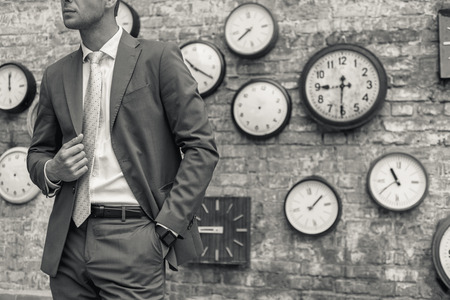 Getting ready for big meeting. Black-white shot of businessman wearing suit and holding his hand in pocket with wall and clock on it in backgroundの写真素材