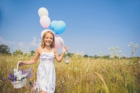 Carefree girl is walking on meadow and smiling. She is holding balloons and basket of flowers. Child is looking at camera with happinessの写真素材