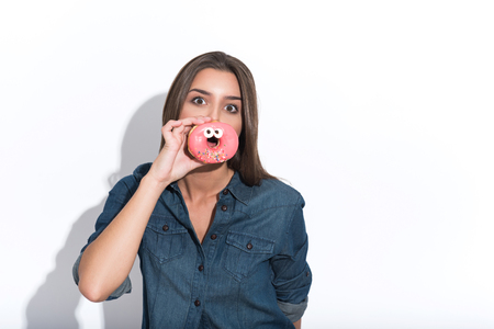 Cute girl is having fun with doughnut. She is covering her mouth with it and looking forward playfully. Isolated and copy space in right sideの写真素材