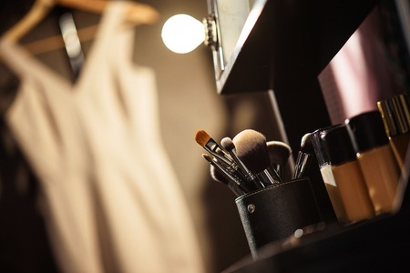Close up of make-up brushes and foundation near mirror backstage. Elegant dress hanging on...