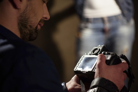 Male photographer is looking at photos in camera with satisfaction. Woman is posing on background in...
