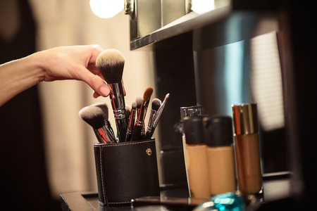 Close up of female hand taking powder brush from set. Various foundations on table near mirror