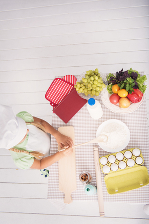 Top view of little girl baking in kitchen. She is holding wooden spoon and taking flour from plate. Kid is standing near table with foodの写真素材