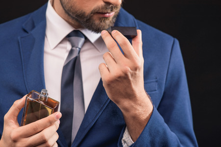 Self-confident young businessman is enjoying smell of masculine perfume. He is standing and holding...