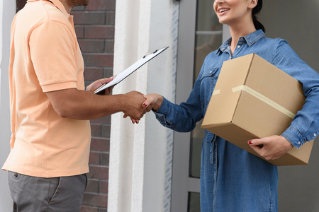 Thank you for timely delivery. Cropped shot of beautiful young woman holding cardboard box and smiling while shaking hands with young delivery manの写真素材