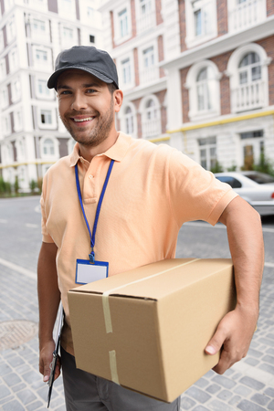 Your parcel is safe with us. Smiling deliveryman holding box while standing on street and looking at cameraの写真素材