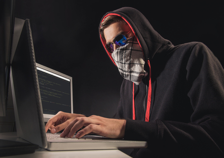 Professional male hacker stealing data from computer. He is sitting at desk in mask and glassesの写真素材