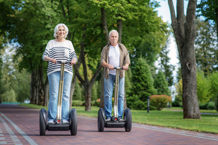 Happy mature man and woman are driving electric scooter in nature. They are standing and smilingの写真素材