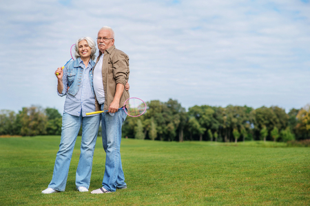 Happy mature man and woman are resting after game in nature. They are standing and holding tennis racks. Husband and wife are embracing and smilingの写真素材