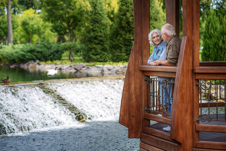 Happy old loving couple is dating in nature. They are standing near lake and talking. Man and woman are smilingの写真素材