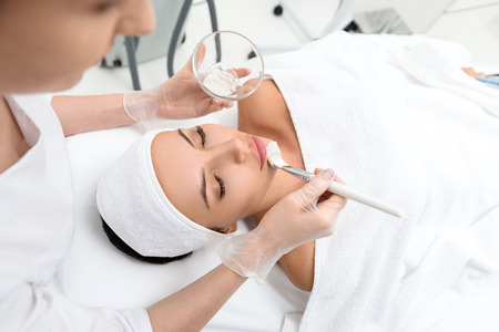 Experienced cosmetician is applying healthy clay on female chin. Girl is lying and relaxing. Her eyes are closed with pleasureの写真素材