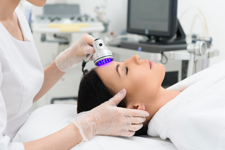 Relaxed young woman is enjoying laser facial massage at medical cosmetology spa salon. She is lying and restingの写真素材