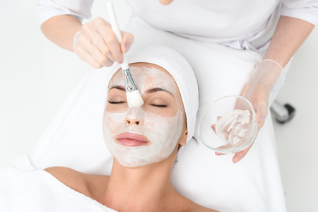 Top view close-up of cosmetologist arms applying cream on female face. She is holding brush and bowl. Young woman is lying and resting. Her eyes are closed with pleasureの写真素材