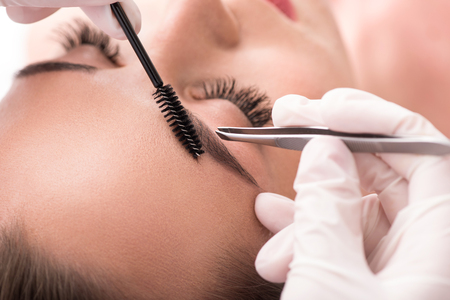 Close up of beautician hands tweezing female eyebrows and combing it by brushの写真素材