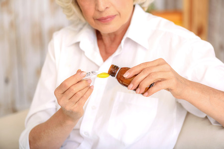 Sick old woman is pouring cough syrup into measure spoon with concentration. She is sitting on sofa at homeの写真素材